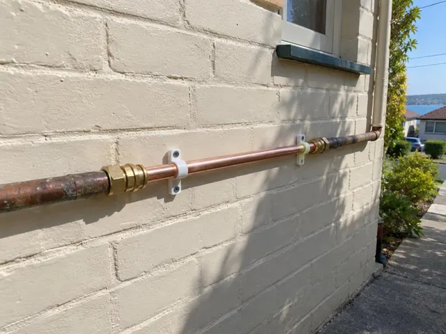 Repaired copper water pipe mounted on a painted brick exterior wall beside a window in Brighton-Le-Sands